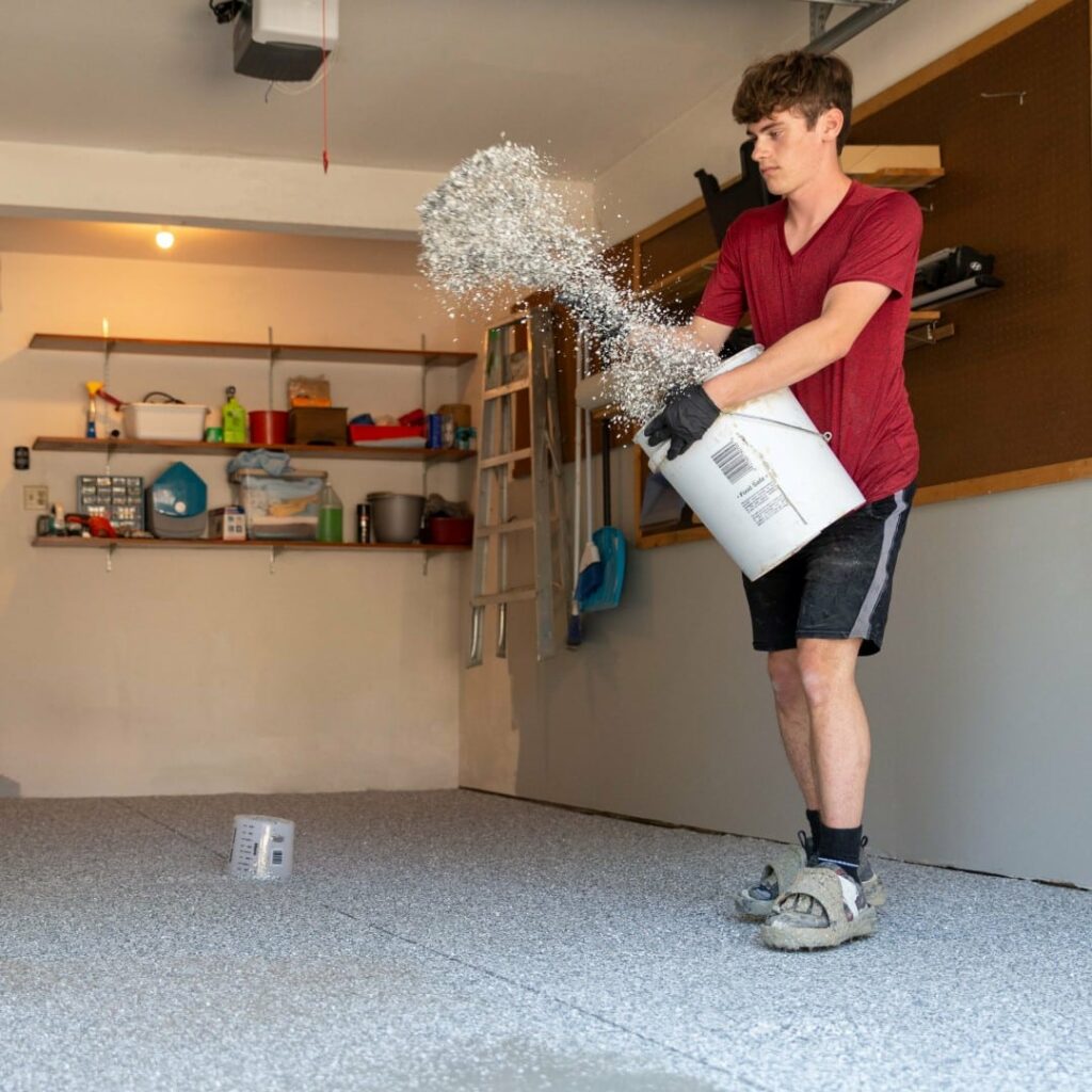 A person in a red shirt is spreading gravel from a bucket onto a garage floor, with shelves and a ladder in the background.