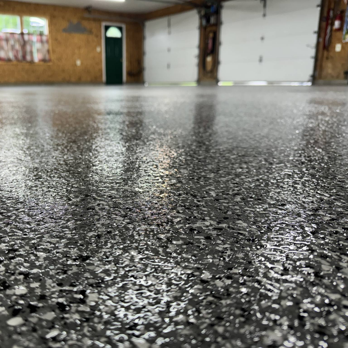 Close-up of a smooth, speckled garage floor under soft lighting, with large closed garage doors and wooden walls in the background.