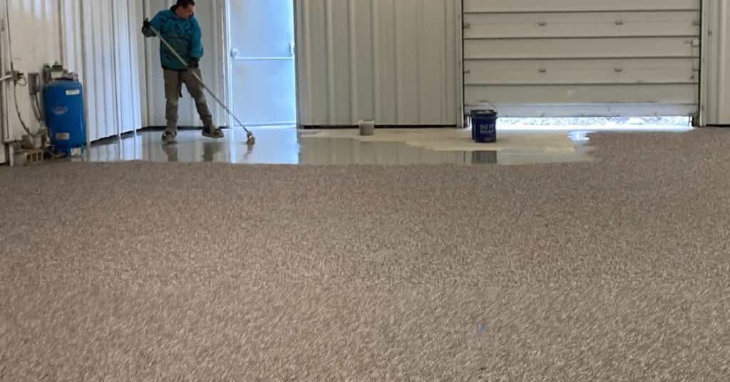 A person applies an epoxy coating to a large indoor floor. Nearby, paint cans and equipment are visible against a metal wall.