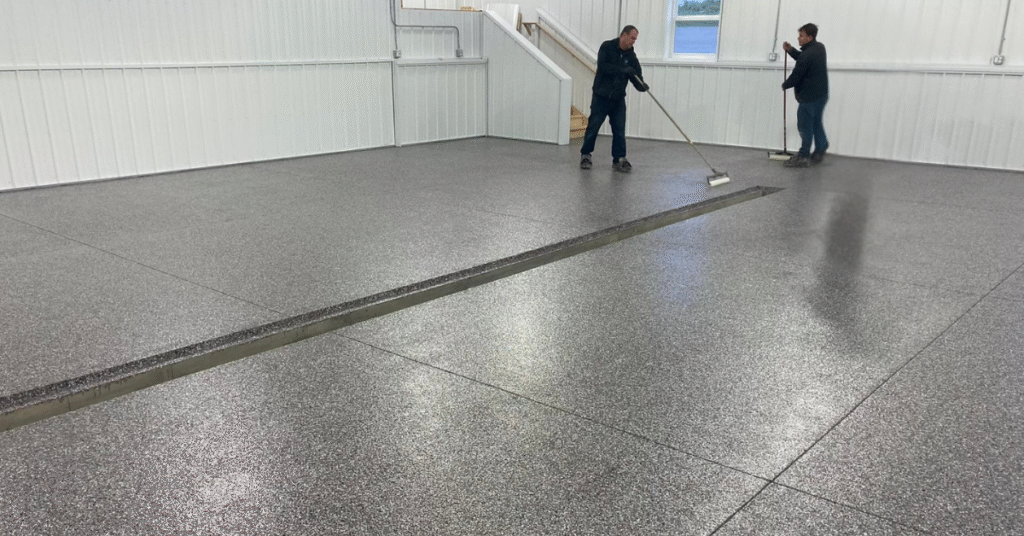 Two people smoothing a newly finished, speckled concrete floor in a large, empty, white-walled room with a staircase and windows.