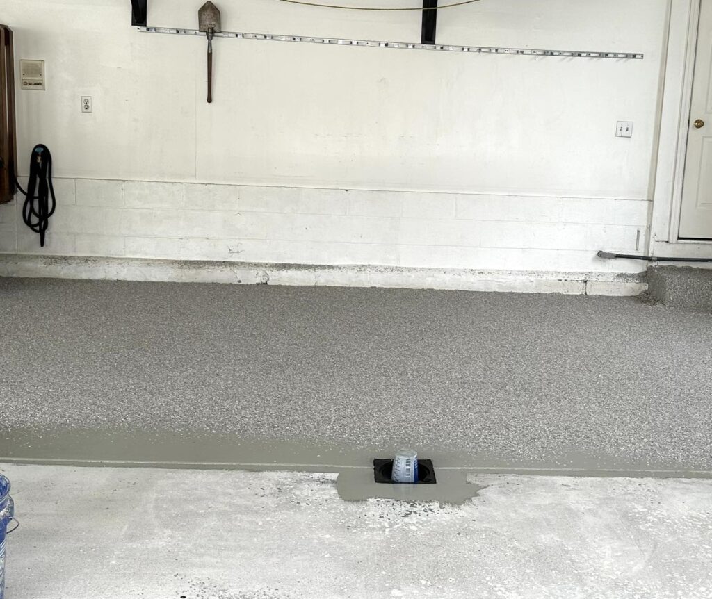 A freshly resurfaced garage floor with exposed wall tools, a door, and a drainage hole in a clean, organized space.