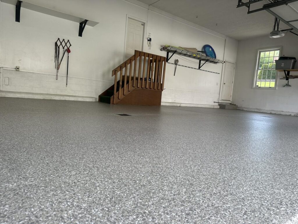 Spacious garage with speckled epoxy floor, wooden stairs, wall shelves, and gardening tools. Closed door and window provide natural light.