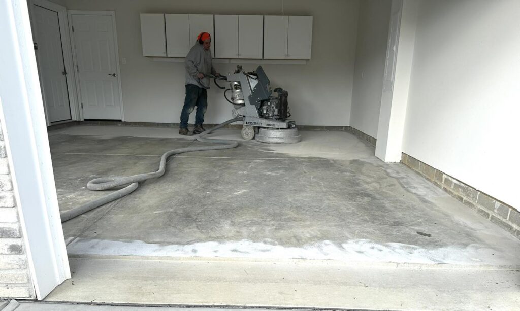 Person grinding a garage floor with a large machine. Gray, smooth concrete surface, with cabinets and a door in the background.