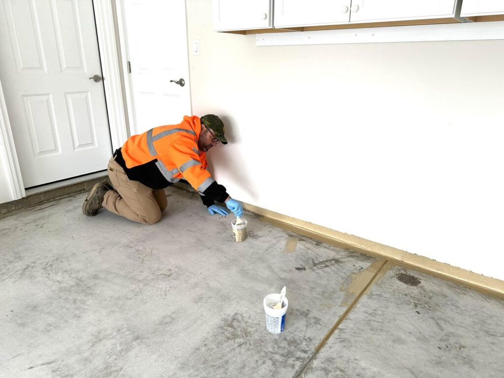 A person in an orange jacket applies filler to a garage floor crack using a plastic spoon, with containers nearby.