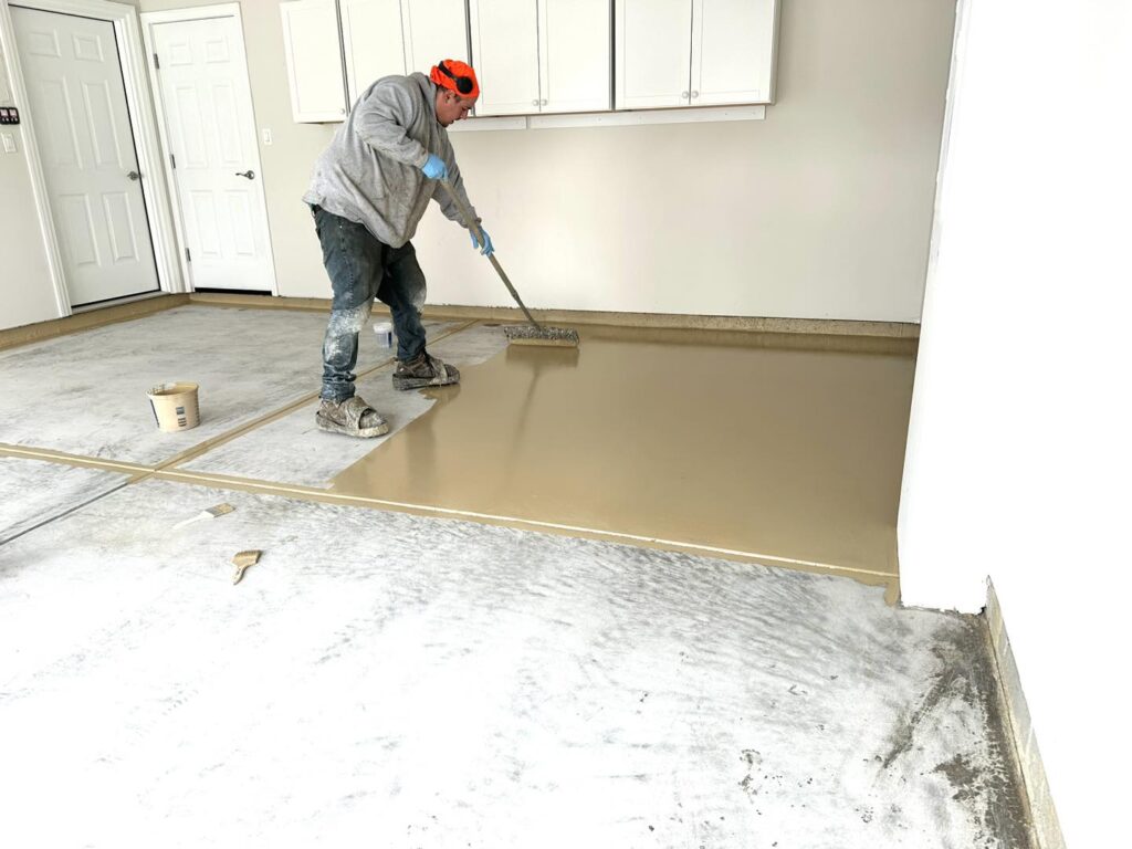 A person in protective gear applies epoxy on a garage floor with a rake tool. White cabinets are mounted on the wall.