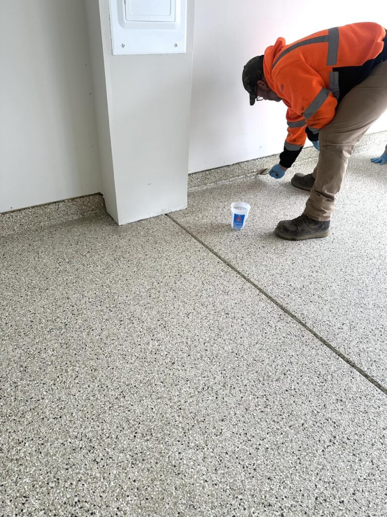 A person in an orange jacket applies epoxy to a garage floor using a brush, with a cup of paint nearby.
