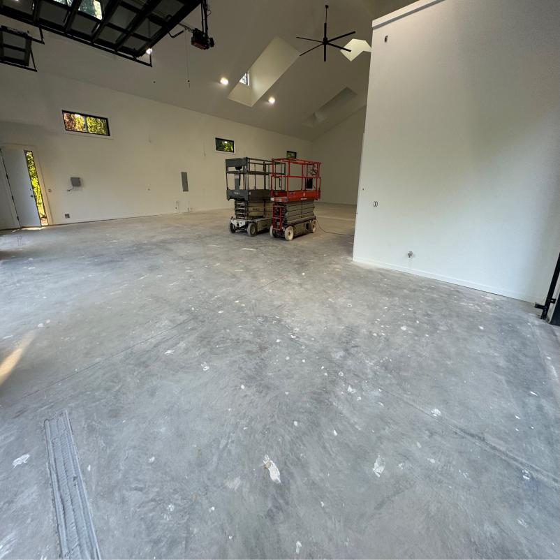 Large, empty indoor space with unfinished concrete floors, white walls, and high ceiling. Two scissor lifts are parked inside. Natural light enters through windows.