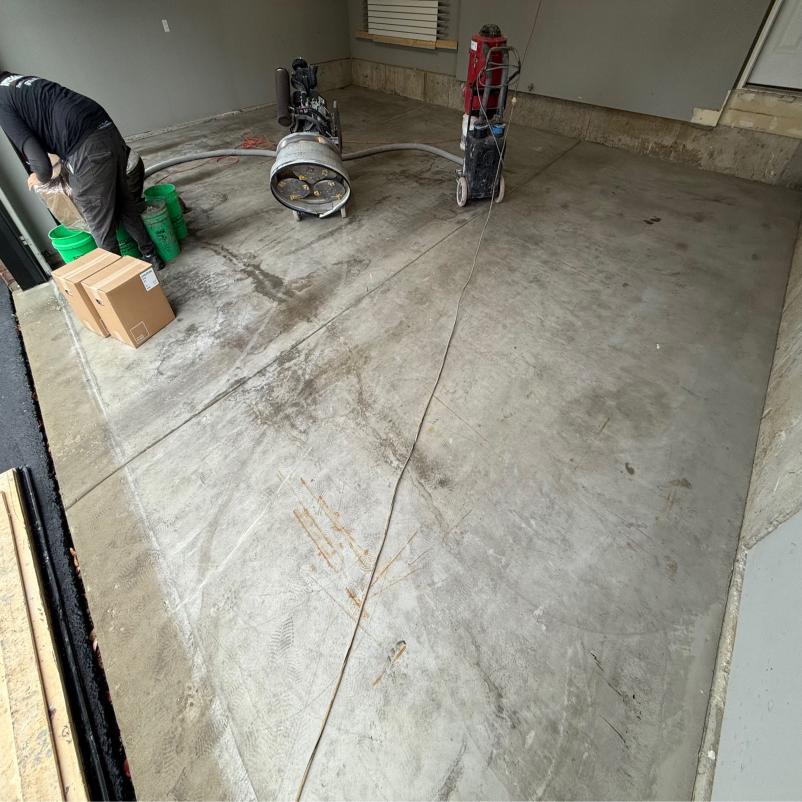 A person working on concrete flooring with polishing equipment, surrounded by green buckets and boxes in a partially closed garage space.